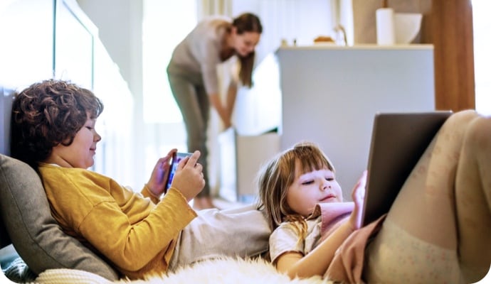 Niños recostados usan tabletas mientras la madre en el fondo organiza la cocina.